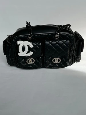 Svart quiltad handväska från Chanel - Säljer en svart quiltad handväska från Chanel i läder med två framfickor och silverfärgade detaljer. Väskan har det klassiska vita CC-logotypen framtill och praktiska fack på sidorna. Perfekt för dig som gillar lyxig och tidlös design.
