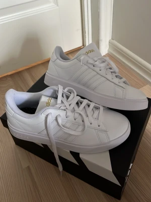 Vita Adidas sneakers med guldlogga - Fräscha vita sneakers från Adidas med klassiska tre ränder på sidorna och guldiga Adidas-loggor på plös och häl. Skorna har en låg profil, vita snören och är tillverkade i slätt syntetmaterial. Perfekta för en clean och stilren look. Fick i julklapp men har aldrig kommit till användning, säljer pga han aldrig lämmna tillbaka dem i tid. Säljer för ordinarie pris. Pris kan diskuteras!! Hör av dig vid mer info! 