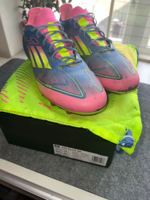 Adidas F50 Elite AG fotbollsskor - Säljer ett par Adidas F50 Elite AG fotbollsskor i en riktigt fet färgkombo av blått, rosa och neongult. Skorna har snörning, låg profil och är tillverkade i lätt syntetmaterial för maxad speed på planen. Lite slitna men går fortfarande att köra med, skopåse ingår och original boxen.