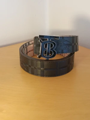 Svart Burberry bälte med rutigt mönster - Stilrent svart bälte från Burberry med klassiskt rutigt mönster och en stor metallspänne med Burberrys ikoniska B-logga. Bältet har en exklusiv känsla och passar perfekt till jeans eller kostymbyxor.fast pris 265kr