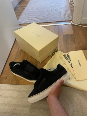 Svarta Axel Arigato sneakers - Stilrena svarta sneakers från Axel Arigato med vit sula och diskret logga på sidan. Skorna har en låg profil och är tillverkade i mocka med minimalistisk design. Perfekta för dig som gillar en clean och modern look. Storlek 41 passar 42