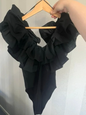  Ny trendig Svart body med volangkrage - Snygg svart body med stor volangkrage runt halsen. Modellen har djup urringning och är tillverkad i ett stretchigt tyg som sitter tight mot kroppen. Perfekt för dig som vill ha något extra till din outfit.