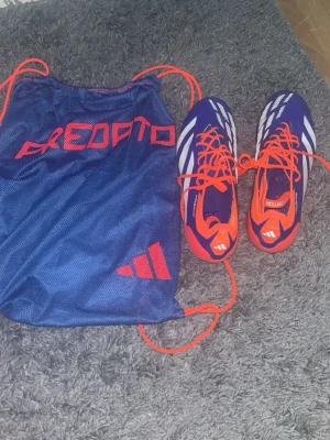 Adidas Elites Predator fotbollsskor blå/orange  - Säljer ett par Adidas Predators Elites fotbollsskor i blått och orange med vita detaljer. Skorna har snörning och är tillverkade i syntetmaterial. Medföljer en blå förvaringspåse med Predator-logga. Perfekta för dig som vill sticka ut på planen! Påsen kommer med på köpet!!!! Köpta för 3000!!!!!
