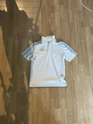 Vit Adidas pikétröja med krage - Snygg vit pikétröja från Adidas med klassiska tre ränder i ljusgrått längs axlarna. Tröjan har korta ärmar, hög krage med knappar och Adidas-logga på bröstet. Perfekt för sport eller träning. Materialet känns lätt och andas bra.