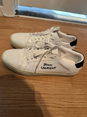 Vita Saint Laurent sneakers i läder - Snygga vita sneakers från Saint Laurent i läder med svart detalj på hälen och handskriven logga på sidan. Klassisk låg modell med vita skosnören och gummisula. Perfekta för en clean och stilren look.