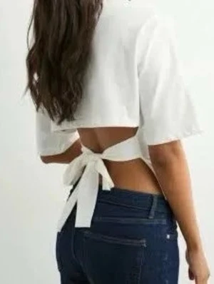 Nelly tröja  - Trendig vit croppad topp med korta ärmar och öppen rygg som knyts med en bred rosett. Perfekt för dig som vill ha en stilren och modern look. Passar snyggt till både jeans och kjol.