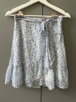 Blommig omlottkjol med volang - Superfin och somrig kjol från VeroModa i ett blått blommönster och volangkant. Kjolen har omlottmodell med knytband i midjan och är tillverkad i ett lätt och luftigt material, perfekt för varma dagar 💕 kjolen är endast använd ett fåtal gånger och säljs pga att den inte kommer till användning 