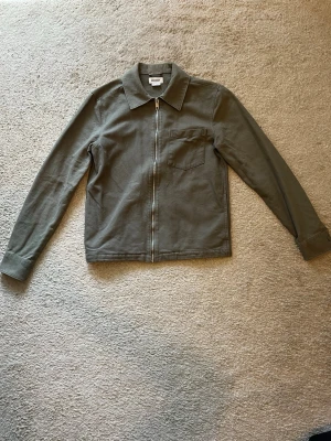 Olivgrön overshirt från Weekday XS - Snygg olivgrön overshirt från Weekday i storlek XS. Jackan har dragkedja framtill, klassisk krage och en bröstficka. Tillverkad i ett mjukt, strukturerat bomullstyg som ger en avslappnad vibe. Perfekt att slänga över en t-shirt.