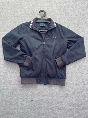 Fred Perry jacka - Klassisk marinblå bomberjacka från Fred Perry med dragkedja framtill, ribbade muddar och krage med vita ränder. Jackan har lång ärm och ikonisk logga broderad på bröstet. Perfekt för en stilren och sportig look.
