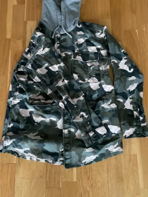 Overshirt i camomönster med huva - Snygg overshirt i grönt, vitt och svart camomönster med grå huva. Två bröstfickor med lock och knappar, samt sidofickor. Långa ärmar med knappslå och avslappnad passform. Perfekt för en streetwear-look.