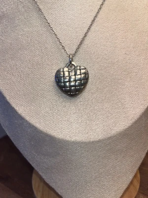 Halsband med hjärtformat silverhänge - Säljer ett halsband med ett hjärtformat hänge i silverfärgad metall. Hänget har ett rutmönster som ger en cool och unik look. Kedjan är tunn och enkel, perfekt för att lyfta din outfit med en personlig touch.