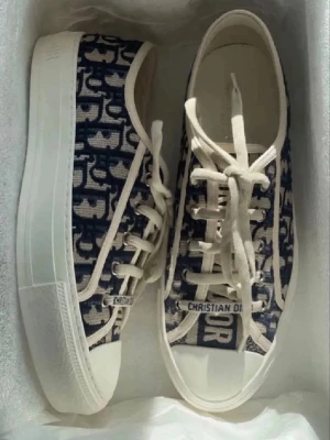 Christian Dior sneakers med monogram - Snygga sneakers från Christian Dior med blått och vitt monogrammönster. Skorna har vit sula, snörning och är tillverkade i textilmaterial med tydliga Dior-detaljer på sidan och tungan. Perfekta för dig som vill sticka ut med lyxig streetstyle.