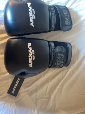 Svarta boxningshandskar Askari - Snygga svarta boxningshandskar från Askari Fight Gear med vit logga och kardborreband. Tillverkade i slitstarkt syntetmaterial, perfekta för träning och sparring. Märket syns tydligt på både handskarna och etiketten.