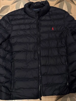 Mainblå  dunjacka från Polo Ralph Lauren storlek XXL - Snygg marinblå dunjacka från Polo Ralph Lauren med klassisk quiltad design och röd broderad logga på bröstet. Jackan har hög krage, dragkedja framtill och två sidofickor med dragkedja. Perfekt för kalla höst och vår dagar. Mycket bra skick. Nypris 3900