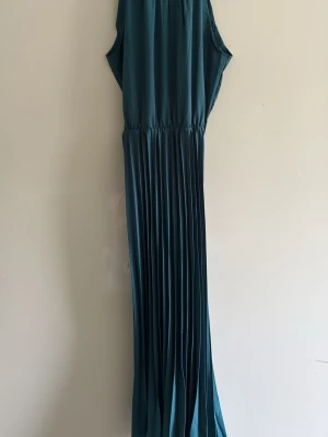 Mörkgrön långklänning i plisserad stil - Säljer en elegant mörkgrön långklänning med plisserad kjol och ärmlös design. Klänningen har en markerad midja och ett mjukt, flytande tyg som ger en snygg siluett. Perfekt för dig som vill ha en stilren och trendig look.