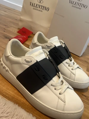 Valentino Garavani vita sneakers - Snygga vita sneakers från Valentino Garavani med bred svart rem över vristen och ikoniska nitar på hälen. Skorna är i läder med vita snören och har en chunky sula för en modern look. Perfekta för dig som vill sticka ut med stilrena detaljer.