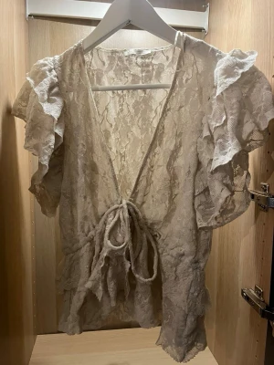 Vit spetsblus med volanger - Romantisk vit blus i spets med djup V-ringning och knytband i midjan. Blusen har korta volangärmar och volangdetaljer längs nederkanten. Perfekt för dig som gillar en feminin och bohemisk stil. Materialet är genombruten spets som ger en luftig känsla. Toppen kan se beige ut på bilden men i verkligheten är toppen en krämig vit.