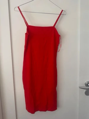 Röd/orange ärmlös klänning med smala axelband - En stilren röd/orange klänning med smala axelband och rak passform. Klänningen är ärmlös och har en enkel, elegant design som passar perfekt för dig som gillar minimalistisk stil. Materialet känns lätt och bekvämt mot huden.