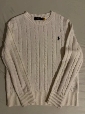 Vit kabelstickad tröja Polo Ralph Lauren - Vit kabelstickad tröja från Polo Ralph Lauren med rund halsringning och broderad marinblå logga på bröstet. Tröjan har långa ärmar och breda ribbade muddar vid ärmslut och nederkant. Klassisk och tidlös design i mjukt material.