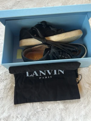 Svarta Lanvin sneakers med lackdetalj - Säljer ett par svarta sneakers från Lanvin med coola lackade tår och klassisk vit sula. Skorna har svarta skosnören och är tillverkade i en mix av textil och mocka. Följer även med ett par läder sko snören. Kommer i originalbox och med dustbag från Lanvin Paris.