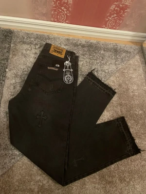 Chrome Hearts svarta jeans med kors - Svarta jeans från Chrome Hearts med broderade korsdetaljer på benen och råa, fransiga benslut. Klassisk femficksmodell med läderpatch bak i midjan och tydliga logodetaljer. Snygg streetstil och unika detaljer som sticker ut.