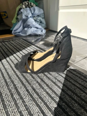 Svarta kilklackssandaler - Snygga svarta sandaler med kilklack från Ally. Skorna har öppen tå, smala remmar över foten och runt ankeln samt spänne för justering. Materialet ser ut att vara mockaimitation som ger en stilren look. Perfekta för dig som vill ha både höjd och komfort.