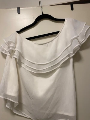 Vit offshoulder blus med volang från Bubbleroom - Superfin vit blus med offshoulder-modell och dubbla volangdetaljer upptill. Luftigt tyg som ger en romantisk vibe, perfekt till jeans eller kjol. Ärmlös på ena sidan och lös ärm på andra för en cool asymmetrisk look.