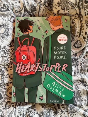 Heartstopper Bok 1 av Alice Oseman - Säljer 'Heartstopper Bok 1' av Alice Oseman, en grafisk roman på svenska. Omslaget är färgglatt med illustration av två killar i skoluniform, en med röd ryggsäck och en med grön axelväska. Boken är mjukband och har ett modernt, ungdomligt uttryck.