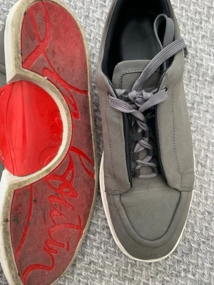 Gråa Louboutin sneakers med röd sula - Stilrena grå sneakers från Christian Louboutin med ikonisk röd sula och diskret Paris-detalj på innersulan. Skorna har en minimalistisk design, rund tå och grå snörning. Ovandelen är i mockaliknande material och sulan är vit med röd undersida.