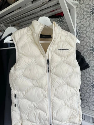 Vit dunväst från Peak Performance - Snygg vit dunväst från Peak Performance med hög krage och dragkedja framtill. Västen har två fickor med dragkedja och ett quiltat mönster som ger en modern look. Perfekt för lager-på-lager-stil under kyliga dagar. Västen är lite missfärgad därefor så billigt pris 