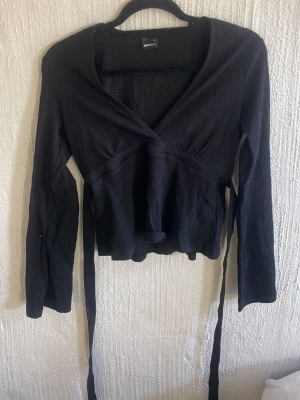 Svart omlottopp från Gina Tricot - Svart omlottopp från Gina Tricot med långa ärmar och knytband i midjan. Toppen har v-ringning och är tillverkad i ett lätt, strukturerat tyg som ger en snygg och trendig look.
