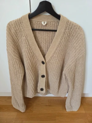 Beige stickad kofta med knappar - Mysig beige stickad kofta med tre stora svarta knappar framtill. Koftan har en djup V-ringning och är gjord i ett grovstickat material som känns både trendigt och bekvämt. Perfekt att slänga på sig över en t-shirt eller linne.