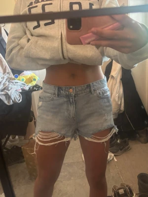 Ljusblå denimshorts med slitningar - Säljer ett par ljusblå denimshorts med råa kanter och slitna detaljer framtill. Shortsen har fem fickor, knapp och dragkedja samt klassisk jeanslook. Perfekta för sommaren och ger en avslappnad vibe.