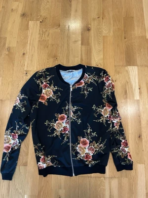 Happy Holly Svart bomberjacka stl 40/42 med blommigt mönster - Snygg svart bomberjacka med färgglatt blommönster i rosa, orange och beige toner. Jackan har dragkedja framtill, ribbade muddar och rundad krage. Perfekt för dig som vill sticka ut med en unik och trendig look.