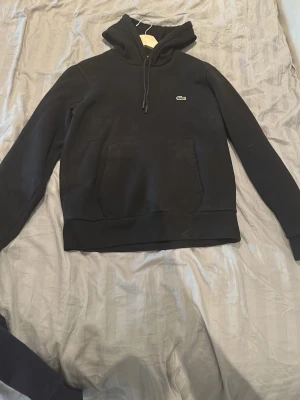 Svart hoodie från Lacoste - Svart hoodie från Lacoste med klassisk krokodillogga på bröstet. Tröjan har huva med dragsko och en stor magficka framtill. Tillverkad i mjukt material som känns skönt mot huden. Perfekt för en avslappnad och stilren look. Storlek S, pris kan diskuteras, om ni har frågor skriv gärna 