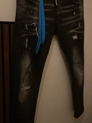 Svarta Dsquared2 jeans med slitningar - Säljer ett par svarta jeans från Dsquared2 med qr kod, coola slitningar och en blå avtagbar detalj med logga. Jeansen har tydliga slitna partier framtill för en trendig look. Perfekta för dig som gillar streetstyle och vill sticka ut.
