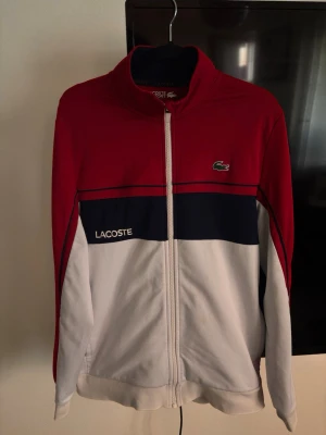 Lacoste hoodie - Snygg hoodie från Lacoste i rött, vitt och marinblått med dragkedja framtill. Klassisk krokodillogga på bröstet och Lacoste-tryck på ena sidan. Mjuk och bekväm med hög krage och långa ärmar. Perfekt för en sportig och stilren look. Enda defekten är att den har en fläck 