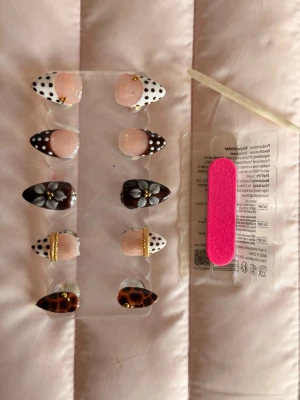 press on nails - Snygga press-on naglar i olika designer: prickiga i svartvitt, bruna med blommor, leopardmönster och gulddetaljer. Inkluderar rosa nagelfil och träpinne för applicering. Naglarna har mandelform och är gjorda av plast.
