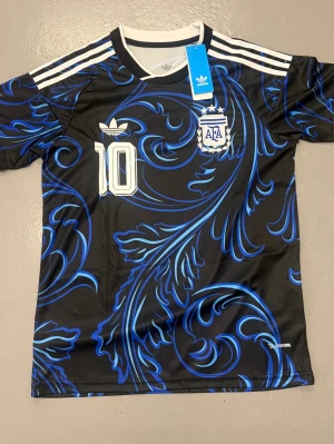 Svart Adidas Argentina Messi tröja - Snygg svart fotbollströja från Adidas med blått mönster, vita detaljer och nummer 10. AFA-logga och Messi-tryck på ryggen. Korta ärmar och klassiska Adidas-ränder på axlarna. Perfekt för dig som älskar fotboll och Messi.