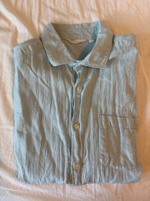 Ljusblå/turkos linneskjorta från Denim Day - Snygg ljusblå skjorta från Denim Day i luftigt linne. Klassisk krage, knäppning framtill och en bröstficka. Perfekt för dig som gillar en avslappnad och stilren look.
