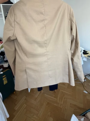 Beige kavaj med två knappar - Stilren beige kavaj med klassisk passform och två knappar framtill. Kavajen har två fickor med lock och en bröstficka. Perfekt för en dressad look och passar till både jeans och kostymbyxor. Materialet känns mjukt och har en lätt struktur.