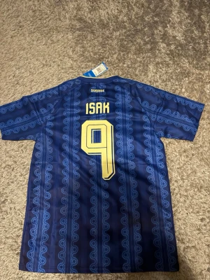 Blå Sverige fotbollströja Adidas - Säljer en blå Sverige fotbollströja från Adidas med Isak och nummer 9 på ryggen. Tröjan har gula detaljer, mönster i blå nyanser och klassiska Adidas-ränder på axlarna. Perfekt för fotbollsfans som vill sticka ut på läktaren.