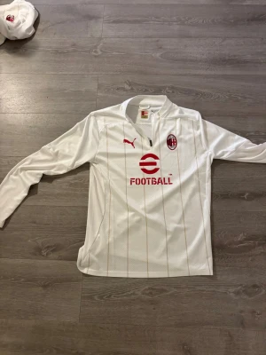 Vit AC Milan träningströja Puma ny och oanvänd  - Helt ny oanvänd snygg långärmad träningströja från AC Milan i vitt med tunna guldiga ränder. Tröjan har halv dragkedja, rött Puma-märke och AC Milan-logga på bröstet samt röd sponsorlogga. Tillverkad i lätt och ventilerande material, perfekt för fotbollsträning.