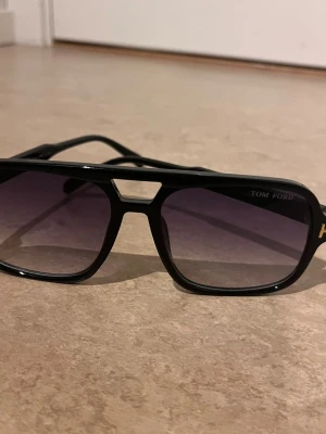 Svarta Tom Ford solglasögon - Snygga svarta solglasögon från Tom Ford med markerad båge och cool detalj över näsryggen. Glasen är mörka och rektangulära, och bågarna har en diskret logga på sidan. Perfekta för dig som vill ha en stilren och trendig look.