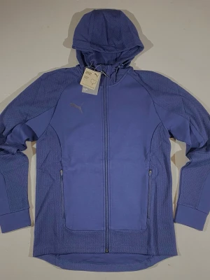 Blå hoodie från Puma med dragkedja - Sportig blå hoodie från Puma med hel dragkedja, huva och två fickor med dragkedja. Materialet är en mix av slät och mesh-liknande textur för extra ventilation. Perfekt för träning eller chill. Puma-logga på bröstet. Finns hos oss i storlek M & S :)