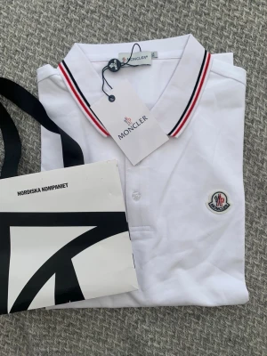 Vit pikétröja från Moncler - Vit pikétröja från Moncler med klassisk krage och knäppning framtill. Kragen har röda och svarta ränder och bröstet pryds av Monclers broderade logotyp. Perfekt för en stilren och sportig look.