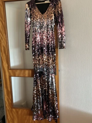 Glittrig långklänning med paljetter - Snygg långklänning från Frock and Frill med glittriga paljetter i guld, silver och rosa toner. Klänningen har v-ringning, lång ärm och figurnära passform. Perfekt för dig som vill sticka ut med en glamorös look.