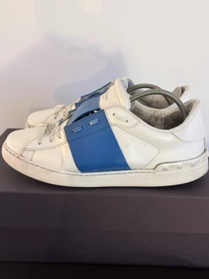 Vita och blå sneakers från Valentino - Snygga vita sneakers från Valentino med en bred blå remsa på sidan och klassiska nitar vid hälen. Skorna har lågt skaft, vita snören och är tillverkade i läder med en stilren och modern design.