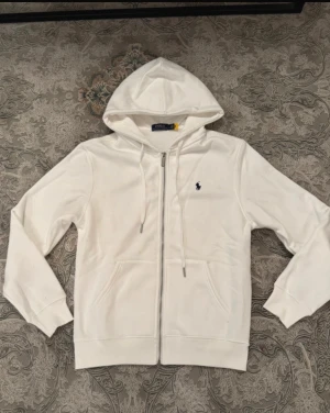 Vit zip hoodie från Polo Ralph Lauren - Snygg vit hoodie med dragkedja från Polo Ralph Lauren. Klassisk design med huva, dragsnören och två fickor framtill. Broderad marinblå logga på bröstet. Perfekt för en clean och stilren look.