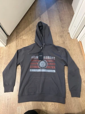 Mörkgrå Saint Laurent hoodie - Helt ny. Snygg mörkgrå hoodie från Saint Laurent med tryck i rött, vitt och grått framtill. Klassisk känguruficka och justerbar huva med dragsko. Perfekt för en avslappnad och trendig stil.